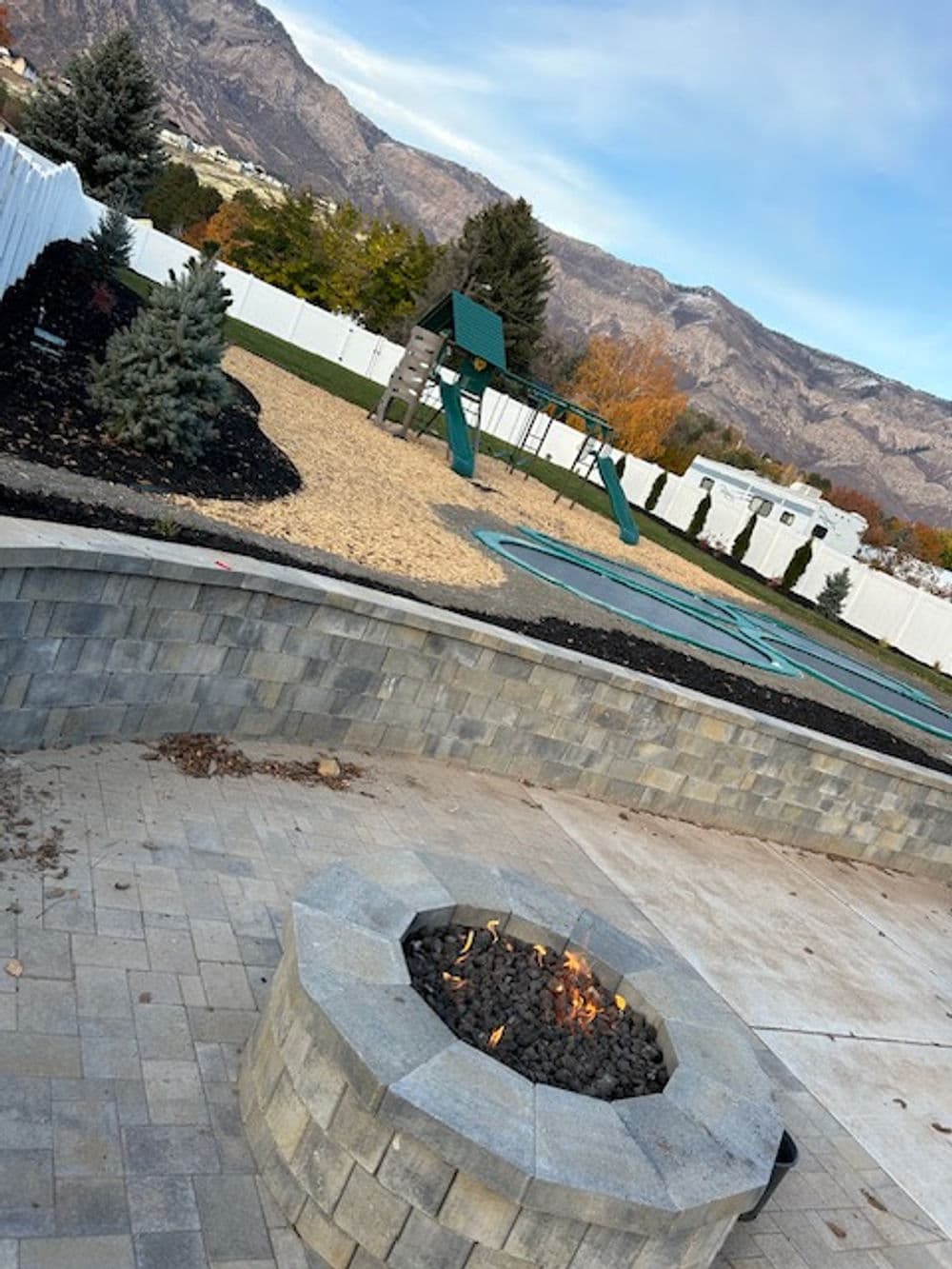 Fire pit in a landscaped backyard with playground and mountain backdrop.