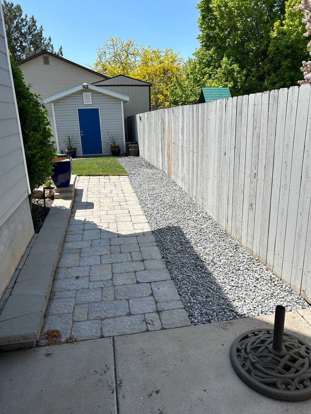 Narrow stone pathway beside wooden fence leading to a blue shed in a sunny backyard.