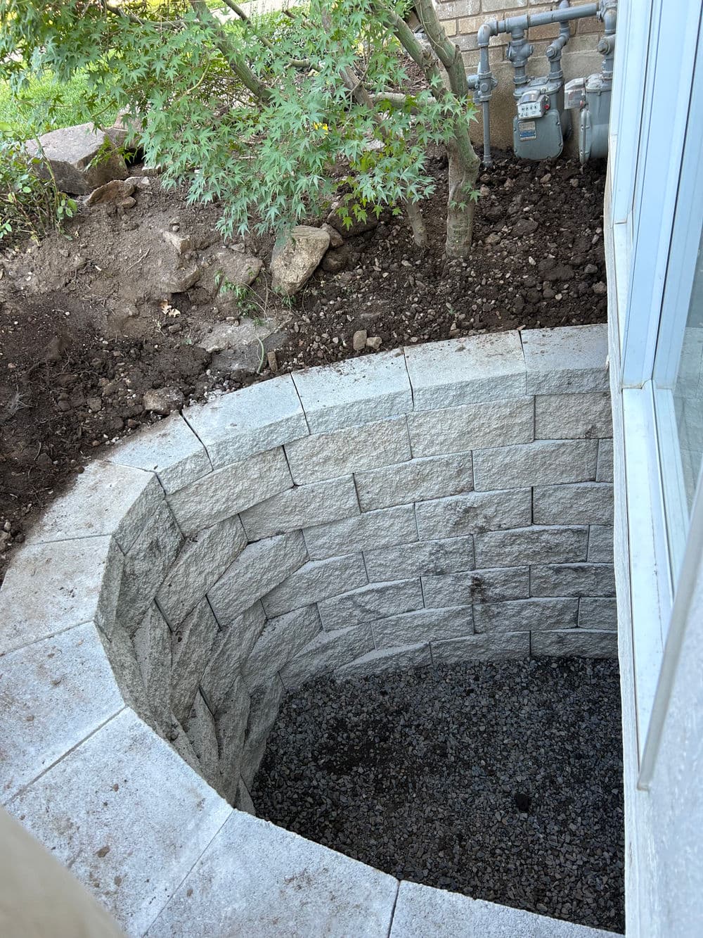 Round stone planter pit with gravel base, surrounded by greenery and utility meters.