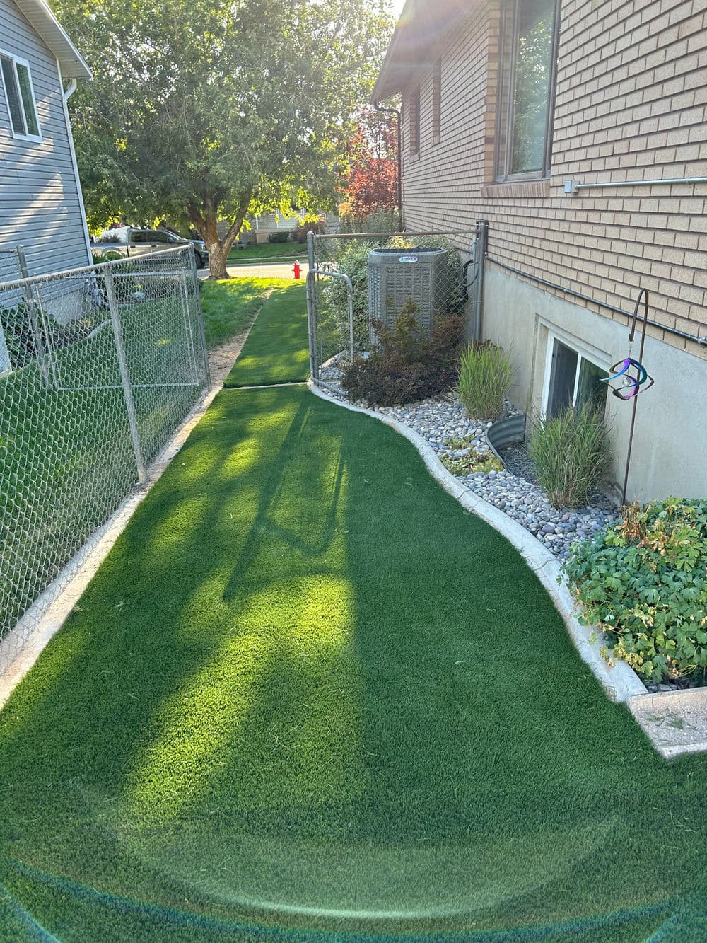 Lush green artificial turf pathway alongside a brick house, bordered by plants and rocks.
