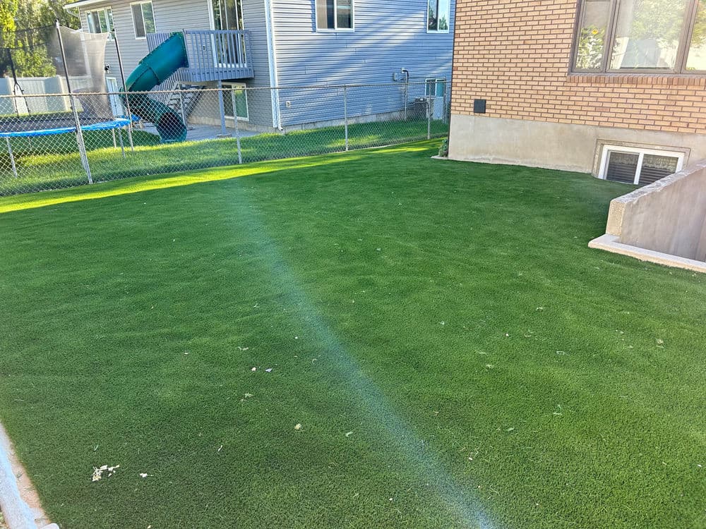 Lush green artificial grass lawn beside a brick house with a playground in the background.