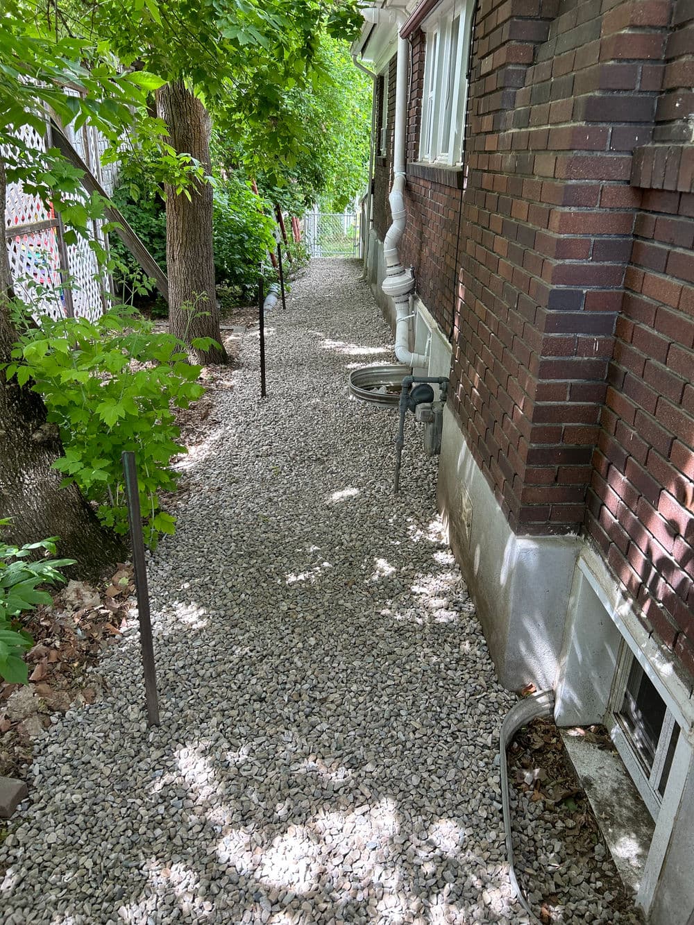 Gravel pathway alongside a brick house, bordered by trees and greenery.